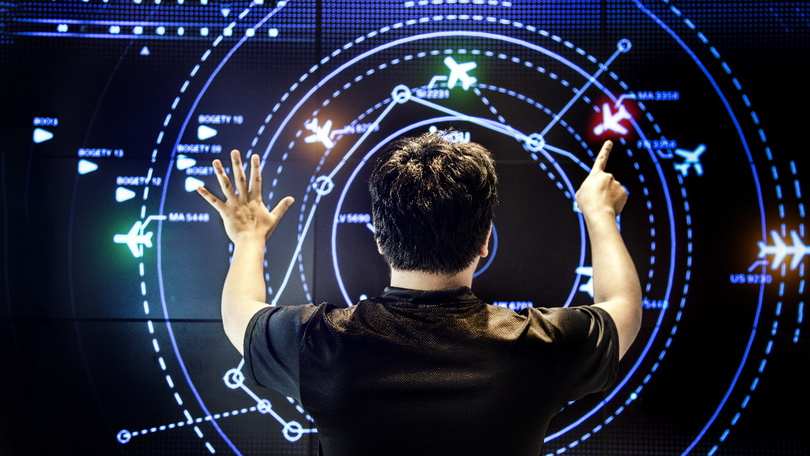 Eine Person steht vor einem digitalen Kontrollpanel. Auf dem Bildschirm werden die Positionen von Flugzeugen dargestellt.
