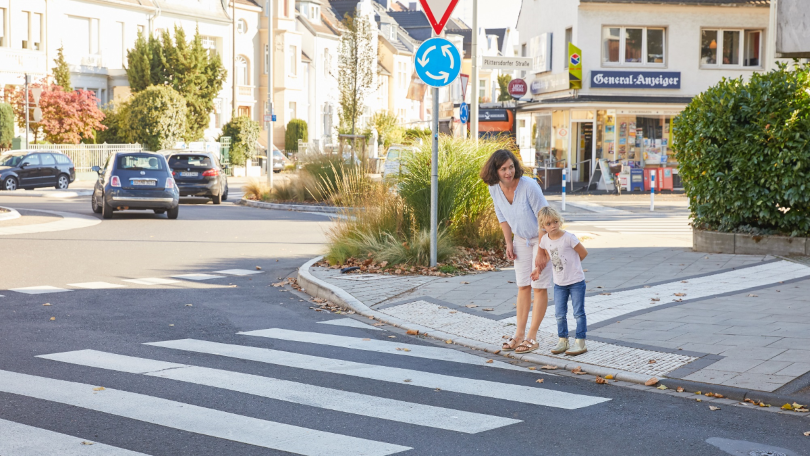 Eine Frau und ein Kind stehen an einem Zebrastreifen und prüfen mit einem Blick nach links, ob die Straße sicher überquert werden kann